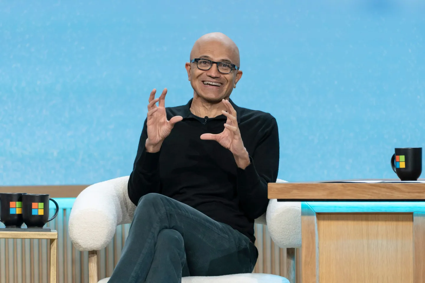Microsoft CEO Satya Nadella sitting on stage at the company&#039;s 50th Anniversary celebrations, wearing a black long-sleeve shirt and black jeans, making a &quot;globe&quot;-like gesture with his two hands.